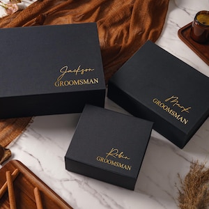Peut inclure: Trois bo&icirc;tes-cadeaux rectangulaires noires de tailles diff&eacute;rentes, chacune orn&eacute;e d'un lettrage dor&eacute;. Les bo&icirc;tes sont personnalis&eacute;es avec les pr&eacute;noms Jackson, Mark et Robin, avec le mot GROOMSMAN imprim&eacute; en majuscules sous chaque pr&eacute;nom.