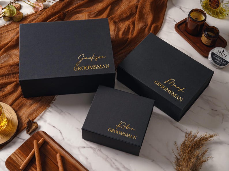 Peut inclure: Trois bo&icirc;tes-cadeaux rectangulaires noires de tailles diff&eacute;rentes, chacune orn&eacute;e d'un lettrage dor&eacute;. Les bo&icirc;tes sont personnalis&eacute;es avec les pr&eacute;noms Jackson, Mark et Robin, avec le mot GROOMSMAN imprim&eacute; en majuscules sous chaque pr&eacute;nom.