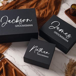 Peut inclure: Trois bo&icirc;tes cadeaux rectangulaires noires avec couvercles &agrave; charni&egrave;res, chacune pr&eacute;sentant des pr&eacute;noms personnalis&eacute;s en &eacute;criture blanche Jackson, James et Nathan, suivis du mot GROOMSMAN en police sans empattement sur le dessus.