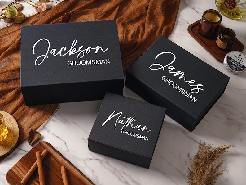 Peut inclure: Trois bo&icirc;tes cadeaux rectangulaires noires avec couvercles &agrave; charni&egrave;res, chacune pr&eacute;sentant des pr&eacute;noms personnalis&eacute;s en &eacute;criture blanche Jackson, James et Nathan, suivis du mot GROOMSMAN en police sans empattement sur le dessus.