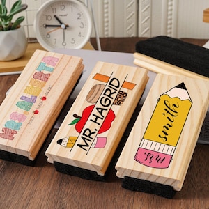 May include: Three wooden dry erase board erasers with teacher-themed designs. One says "Math Teacher" in colorful letters, another says "Mr. Hagrid" with an apple, and the last says "Smith" with a pencil illustration.