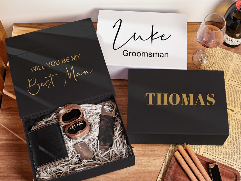 Peut inclure: Un coffret cadeau noir avec le texte dor&eacute; WILL YOU BE MY Best Man contenant une flasque noire, une bo&icirc;te &agrave; bagues en bois et un briquet. &Agrave; c&ocirc;t&eacute; se trouvent une bo&icirc;te blanche avec le texte Luke Groomsman et une bo&icirc;te noire avec le texte dor&eacute; THOMAS.