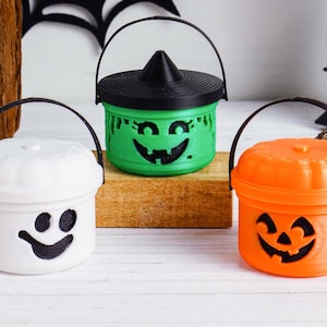 May include: Three Halloween-themed buckets with black handles. One is white with a smiling face, one is green with a witch hat lid and a carved face, and the last is orange with a jack-o'-lantern face. All have black smiling faces.