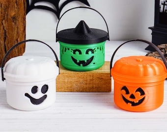 May include: Three Halloween-themed buckets with black handles. One is white with a smiling face, one is green with a witch hat lid and a carved face, and the last is orange with a jack-o'-lantern face. All have black smiling faces.
