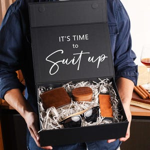 Peut inclure: Une bo&icirc;te cadeau rectangulaire noire avec le texte IT'S TIME to Suit up imprim&eacute; sur le couvercle int&eacute;rieur. La bo&icirc;te contient un portefeuille en cuir marron, un petit r&eacute;cipient en bois, un accessoire en cuir beige et des lunettes noires.