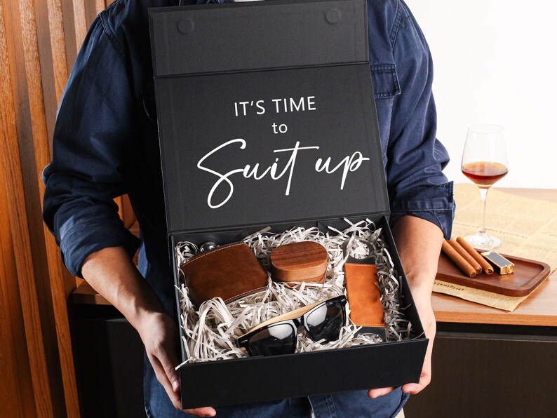 Peut inclure: Une bo&icirc;te cadeau rectangulaire noire avec le texte IT'S TIME to Suit up imprim&eacute; sur le couvercle int&eacute;rieur. La bo&icirc;te contient un portefeuille en cuir marron, un petit r&eacute;cipient en bois, un accessoire en cuir beige et des lunettes noires.