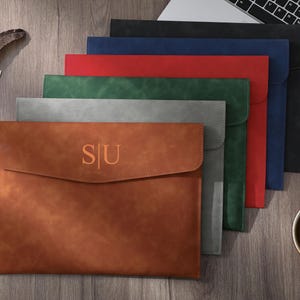Peut inclure: Une pile de pochettes à documents rectangulaires en marron, gris, vert, rouge, bleu et noir. La pochette marron située à l'avant présente les lettres S|U en relief sur le rabat. Chaque pochette a une finition mate et un rabat incurvé.