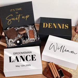 Peut inclure: Trois bo&icirc;tes cadeaux rectangulaires noires et blanches avec des noms et dates personnalis&eacute;s. Une bo&icirc;te noire ouverte contient une flasque marron, des lunettes de soleil et des accessoires. Les bo&icirc;tes portent les textes IT'S TIME TO SUIT UP, DENNIS, LANCE et WILLIAM.