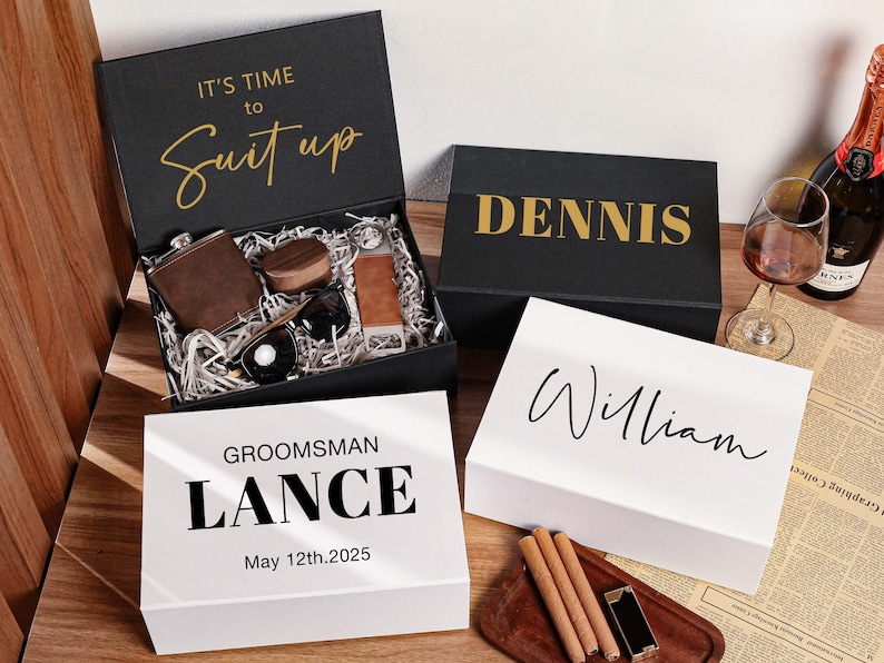 Peut inclure: Trois bo&icirc;tes cadeaux rectangulaires noires et blanches avec des noms et dates personnalis&eacute;s. Une bo&icirc;te noire ouverte contient une flasque marron, des lunettes de soleil et des accessoires. Les bo&icirc;tes portent les textes IT'S TIME TO SUIT UP, DENNIS, LANCE et WILLIAM.