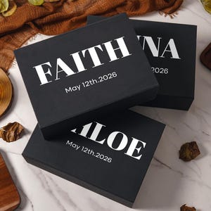 Peut inclure: Trois bo&icirc;tes cadeaux rectangulaires noires avec des inscriptions blanches. La bo&icirc;te sup&eacute;rieure affiche le nom FAITH et la date May 12th.2026. Une autre bo&icirc;te montre le nom ALOE et la date May 12th.2026. La troisi&egrave;me bo&icirc;te montre le nom partiel NA et 2026.