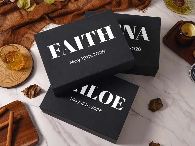 Peut inclure: Trois bo&icirc;tes cadeaux rectangulaires noires avec des inscriptions blanches. La bo&icirc;te sup&eacute;rieure affiche le nom FAITH et la date May 12th.2026. Une autre bo&icirc;te montre le nom ALOE et la date May 12th.2026. La troisi&egrave;me bo&icirc;te montre le nom partiel NA et 2026.