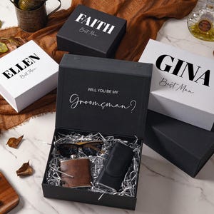 Peut inclure: Une collection de bo&icirc;tes cadeaux noires et blanches avec des pr&eacute;noms personnalis&eacute;s comme FAITH, ELLEN et GINA imprim&eacute;s sur les couvercles. Une bo&icirc;te noire ouverte contient du papier argent&eacute;, une flasque en cuir marron, des lunettes et un &eacute;tui.