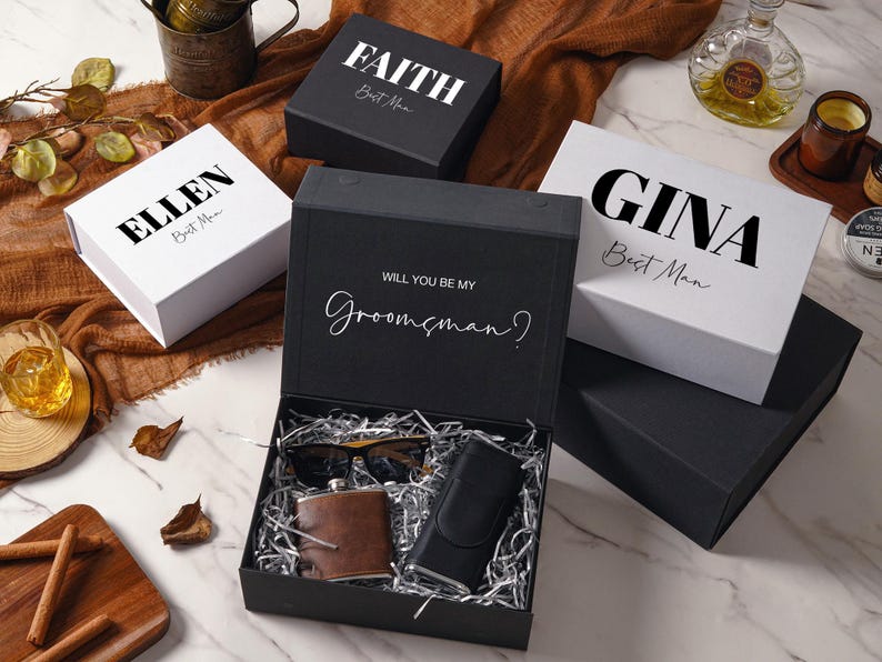 Peut inclure: Une collection de bo&icirc;tes cadeaux noires et blanches avec des pr&eacute;noms personnalis&eacute;s comme FAITH, ELLEN et GINA imprim&eacute;s sur les couvercles. Une bo&icirc;te noire ouverte contient du papier argent&eacute;, une flasque en cuir marron, des lunettes et un &eacute;tui.