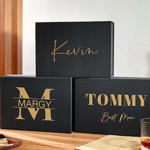 Peut inclure: Trois bo&icirc;tes cadeaux rectangulaires noires avec des inscriptions dor&eacute;es. La bo&icirc;te sup&eacute;rieure porte le pr&eacute;nom Kevin en &eacute;criture cursive. La bo&icirc;te en bas &agrave; gauche affiche un grand M avec le pr&eacute;nom Margy. La bo&icirc;te en bas &agrave; droite indique Tommy Best Man.