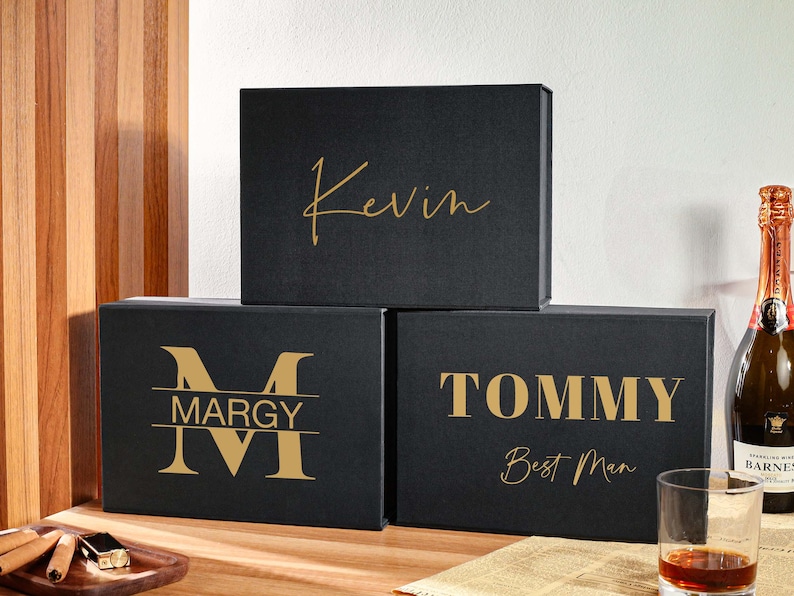 Peut inclure: Trois bo&icirc;tes cadeaux rectangulaires noires avec des inscriptions dor&eacute;es. La bo&icirc;te sup&eacute;rieure porte le pr&eacute;nom Kevin en &eacute;criture cursive. La bo&icirc;te en bas &agrave; gauche affiche un grand M avec le pr&eacute;nom Margy. La bo&icirc;te en bas &agrave; droite indique Tommy Best Man.