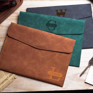 May include: Three rectangular document folders in brown, green, and dark gray. Each folder features a flap closure and a personalized engraving with a large initial and a name, specifically Loren, Gavin, and William, centered on the front.