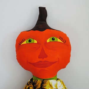 May include: A handmade orange fabric doll with a smiling face, green eyes, and a brown stem. The doll has a yellow and black patterned collar. A whimsical, decorative item.