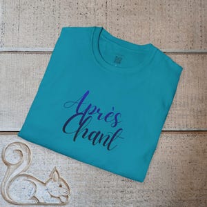 Op de afbeelding: Turquoisekleurig T-shirt met de tekst "Après Chant" in een blauw-paarse gradiënt. Het shirt is opgevouwen op een houten oppervlak, met een decoratief element in de vorm van een eekhoorn in de hoek.