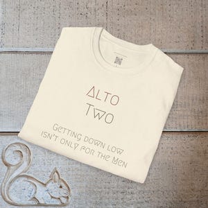 Puede incluir: Camiseta color crema con el texto "ALTO TWO" en rojo y negro, y la frase "Getting down low isn't only for the men." La camiseta se muestra sobre una superficie de madera clara.