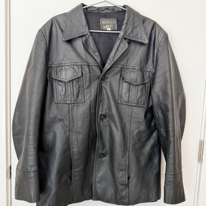 May include: A black leather jacket featuring a pointed collar, a button-front closure, and two chest pockets with flaps. The interior label displays the text ECHTES LEDER. The jacket has long sleeves with cuffs and a smooth, matte finish throughout.