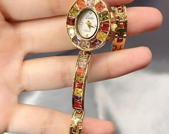 May include: A gold-tone quartz watch featuring an oval white dial with the text Lsvtr. The bezel and bracelet are embellished with square-cut, multi-colored gemstones in shades of red, orange, yellow, and purple, set in a polished metal frame.