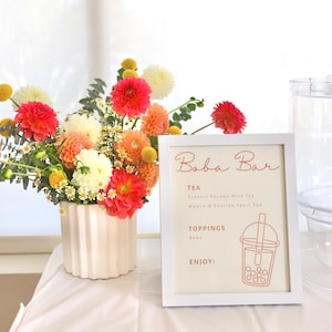 Puede incluir: Un cartel enmarcado en blanco con el texto "Boba Bar" en escritura rosa. El cartel enumera "Té" con las opciones "Té de leche Dolong clásico" y "Té de mango y maracuyá". También enumera "Ingredientes" con la opción "Boba". El cartel tiene un dibujo de línea rosa de una taza de té boba con una pajita.