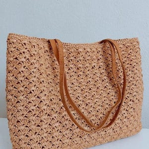 May include: A woven tote bag in a light tan color with brown leather handles. The bag has a textured, open weave design. The handles are long enough to be worn over the shoulder. The bag is sitting on a white surface.