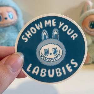 Könnte beinhalten: Ein runder Aufkleber mit dem Text "SHOW ME YOUR LABUBIES" in weißer Schrift auf dunkelblauem Hintergrund. Der Aufkleber zeigt eine Cartoonfigur mit einem hasenähnlichen Gesicht und einem grinsenden Mund. Zwei Plüschtiere sind im Hintergrund zu sehen.