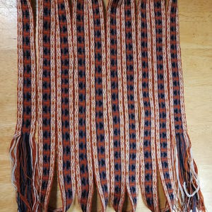 Puede incluir: Siete tiras de tejido tejido con un patrón geométrico en tonos marrones, blancos y negros. Las tiras tienen flecos en los extremos.