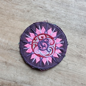 Könnte beinhalten: Ein runder, gestickter Aufnäher mit lila Hintergrund und einem rosa-roten Blumenmuster. Der Aufnäher hat ein detailliertes, stilisiertes Sonnenmuster mit einem zentralen Wirbeldesign. Der Aufnäher hat einen Durchmesser von ca. 7,6 cm.
