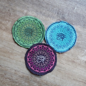 Pode incluir: Três patches circulares bordados em tons de verde, azul e roxo. Cada patch apresenta um design de Olho de Hórus no centro, rodeado por um padrão simétrico detalhado. Os patches são exibidos em uma superfície de madeira.