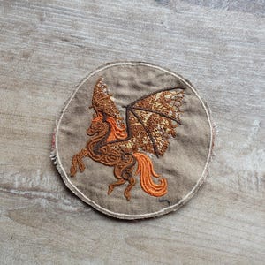 Puede incluir: Parche bordado circular que representa un caballo alado en tonos marrón y naranja. El parche presenta un diseño detallado de la criatura mítica con alas extendidas y una cola ondeante, sobre una tela beige. Adecuado para ropa o accesorios.