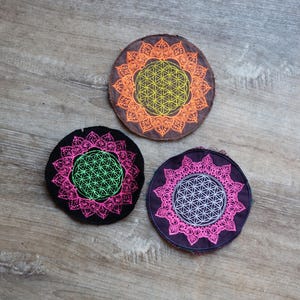 May include: Three round coasters with a floral design. One coaster is orange and yellow, another is black and pink, and the third is purple and pink. The coasters are placed on a wooden surface.