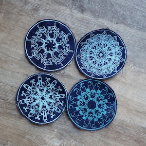 May include: Four navy blue coasters with intricate, light blue embroidered mandala designs. Each coaster features a different symmetrical pattern, creating a set with a cohesive aesthetic. The coasters are round and sit on a wooden surface.