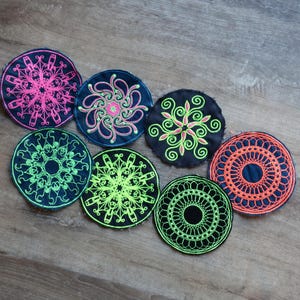 Puede incluir: Colección de posavasos circulares bordados en varios colores, incluyendo azul marino, negro y naranja. Cada posavasos presenta un diseño único e intrincado en colores contrastantes como rosa, verde y naranja. Los posavasos están dispuestos sobre una superficie de madera.