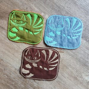 May include: Three square embroidered patches featuring a grinning cat design. One patch is green with lime green embroidery, another is blue with light blue embroidery, and the third is brown with beige embroidery. The patches are on a wooden surface.