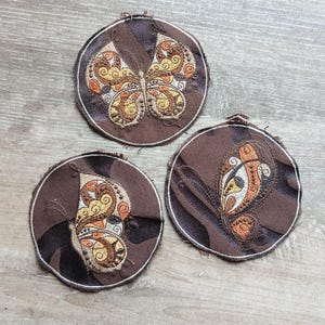 Pode incluir: Três bases para copos redondas de tecido castanho com bordados de borboletas. As borboletas apresentam detalhes em laranja, amarelo e branco. As bases têm uma borda desfiada, sobre um fundo de madeira.