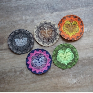 Könnte beinhalten: Fünf farbige, runde Untersetzer mit gestickten Sonnen-Gesichts-Designs. Die Untersetzer sind in Grautönen, Braun, Orange, Rosa und Grün gehalten. Jeder Untersetzer hat ein detailliertes Sonnen-Gesichts-Design mit einem dekorativen Rand.