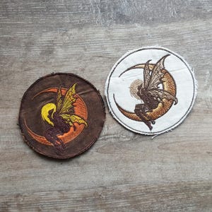 May include: Two round embroidered patches featuring a fairy sitting on a crescent moon. One patch is brown with yellow, orange, and brown details. The other is white with similar colors. The patches are on a wooden surface.