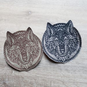 May include: Two circular embroidered patches featuring wolf heads. One is brown with a detailed design, the other navy blue. Both patches have intricate patterns and are suitable for clothing or accessories.