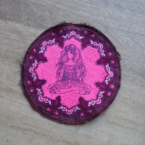 May include: A round, embroidered patch featuring a seated figure in a yoga pose. The patch has a pink center with a detailed purple figure and a decorative border. The outer edge is a darker purple with a frayed texture.