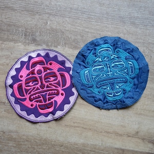 Peut inclure: Deux écussons ronds brodés avec des motifs d'inspiration amérindienne. L'un est violet avec des détails roses, l'autre est bleu avec des accents turquoise. Les deux présentent des visages stylisés et des coutures complexes, parfaits pour coudre sur des vêtements ou des accessoires.