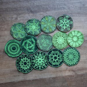 Puede incluir: Una colección de posavasos redondos bordados con intrincados diseños verdes brillantes. Los posavasos presentan varios patrones, incluyendo motivos florales, geométricos y tipo mandala. Los posavasos están dispuestos sobre una superficie de madera.