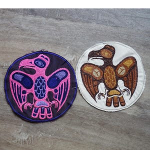 Op de afbeelding: Twee ronde, geborduurde patches met adelaarsontwerpen. Eén patch heeft een roze adelaar op een donkerblauwe achtergrond, de andere een bruine adelaar op een witte achtergrond. Geschikt voor kleding.