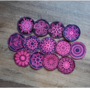 Könnte beinhalten: Eine Sammlung runder, gestickter Untersetzer mit komplizierten rosa Mandala-Designs auf dunkelblauem Hintergrund. Die Untersetzer sind auf einer Holzoberfläche angeordnet und zeigen verschiedene Blumen- und geometrische Muster.