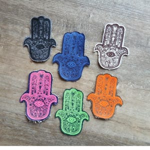 May include: A collection of six embroidered Hamsa hand patches in various colors: black, blue, pink, green, and orange. Each patch features an eye design in the center and intricate detailing. The patches are displayed on a wooden surface.