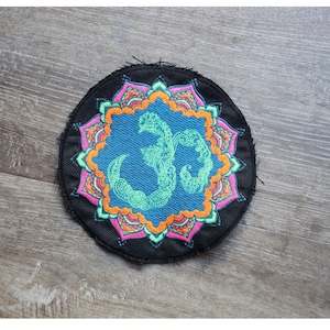 Può includere: Una toppa rotonda ricamata nera con un centro blu con un simbolo Om verde. Il simbolo è circondato da un design floreale dettagliato e colorato nei toni dell'arancione, del rosa e del verde. La toppa ha un aspetto strutturato.