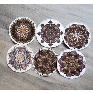 May include: Six round coasters with intricate mandala designs in shades of brown, gold, and white. The coasters are arranged on a wooden surface, showcasing their decorative patterns. These coasters are ideal for protecting surfaces from drink rings.