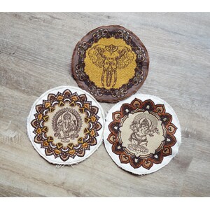 May include: Three round coasters with embroidered designs. One features an elephant, another depicts a deity, and the third showcases a couple. The coasters have a white border and brown and gold accents, set against a wooden background.
