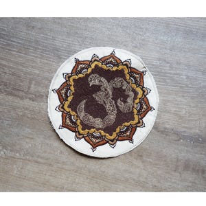 May include: Round, off-white coaster with a brown and gold mandala design. The center features a brown Om symbol with intricate gold detailing. The coaster is likely made of fabric or a similar material.